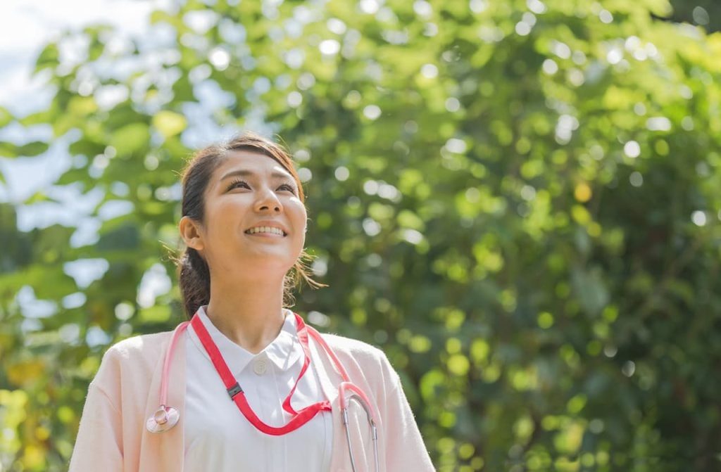 白衣にピンクのカーディガンを着た看護師が、天気のいい屋外で微笑んでいる